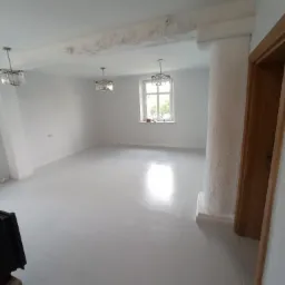 Białe, przestronne wnętrze po remoncie z nową, błyszczącą podłogą z płytek, widoczną kolumną dekoracyjną ze strukturą i trzema wiszącymi lampami z kryształkami. Na suficie widoczne ślady po...