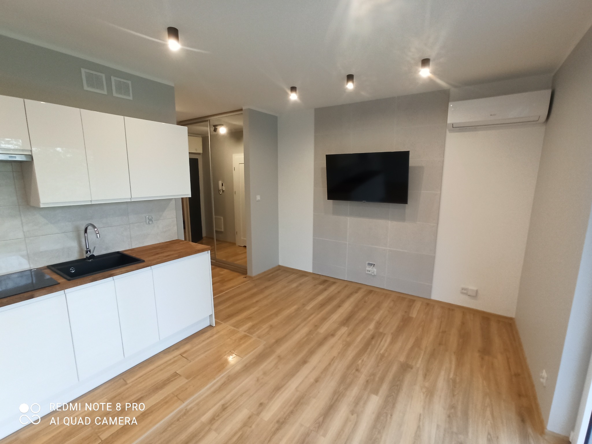 Jasne wnętrze po remoncie: minimalistyczna kuchnia z białymi szafkami i drewnianym blatem, drewnopodobna podłoga, szara ściana z telewizorem i klimatyzacją, punktowe oświetlenie sufitowe.