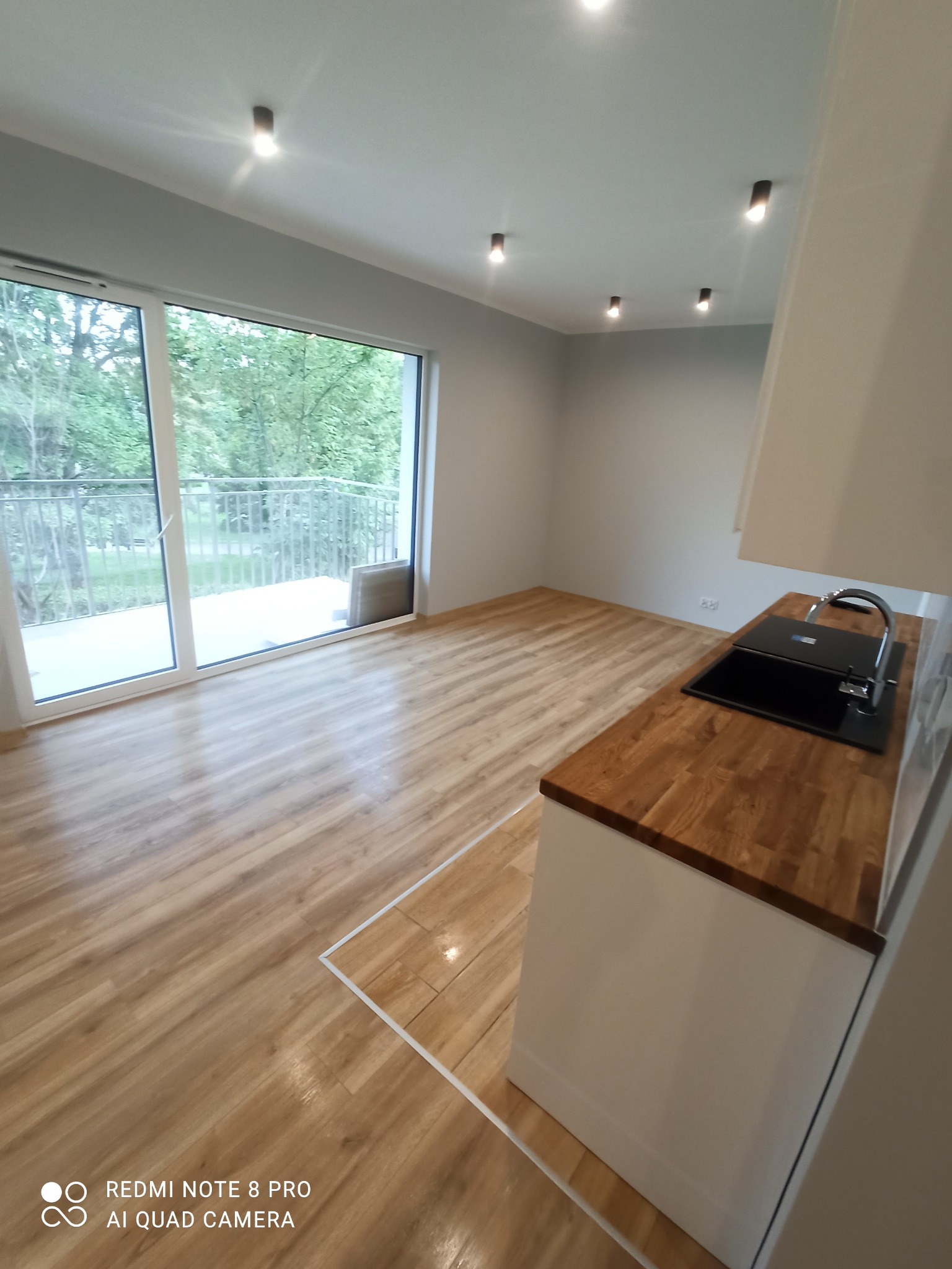 Nowoczesna kuchnia w jasnych kolorach z drewnianym blatem, widok na balkon i zieleń za oknem, oświetlenie punktowe na suficie.