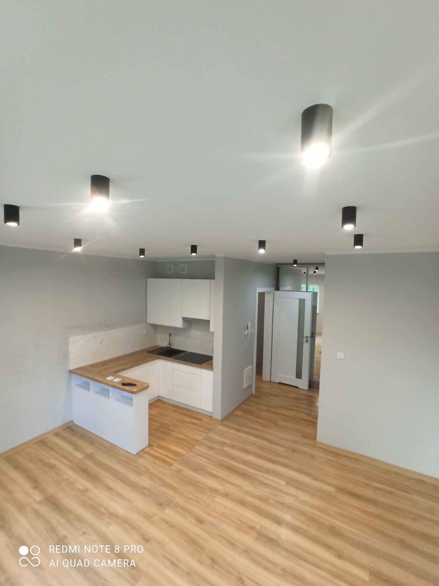 Widok na nowocześnie wykończone wnętrze: jasna kuchnia z drewnianym blatem i czarnym zlewem, parkiet w odcieniach beżu, szare ściany i sufit z punktowym oświetleniem LED.