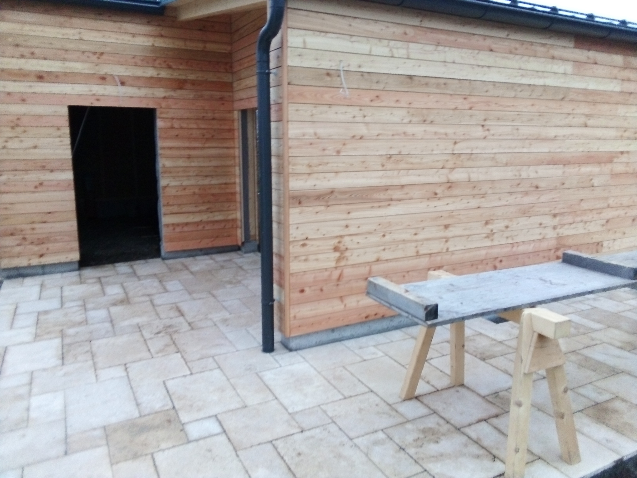 Świeżo położona, pozioma boazeria z jasnego drewna na ścianie budynku z otworami na drzwi, widoczne rusztowanie i kostka brukowa na posadzce.