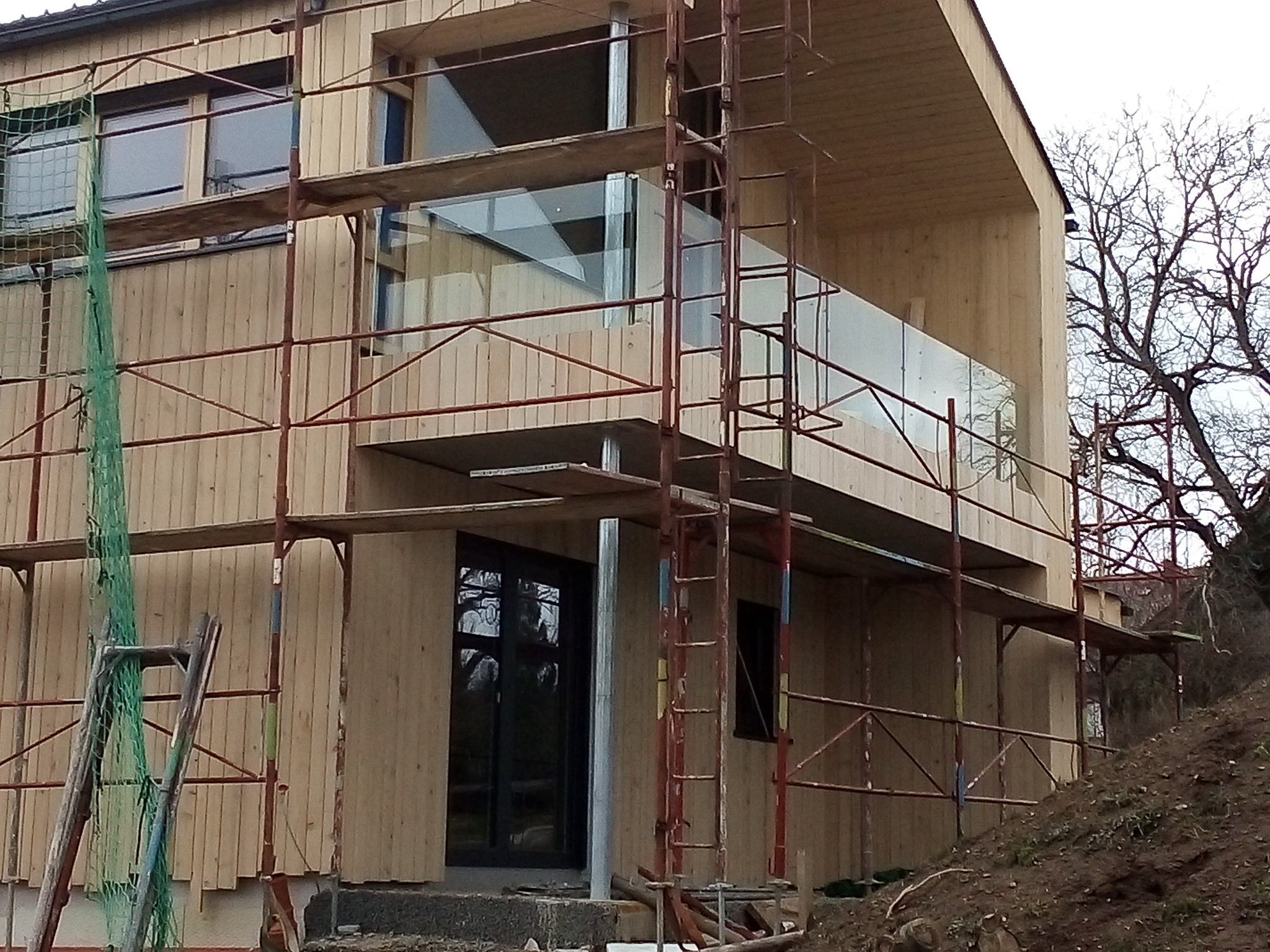 Montaż elewacji drewnianej z balkonem i szklaną balustradą na budynku w trakcie budowy, widoczne rusztowania i drabina.