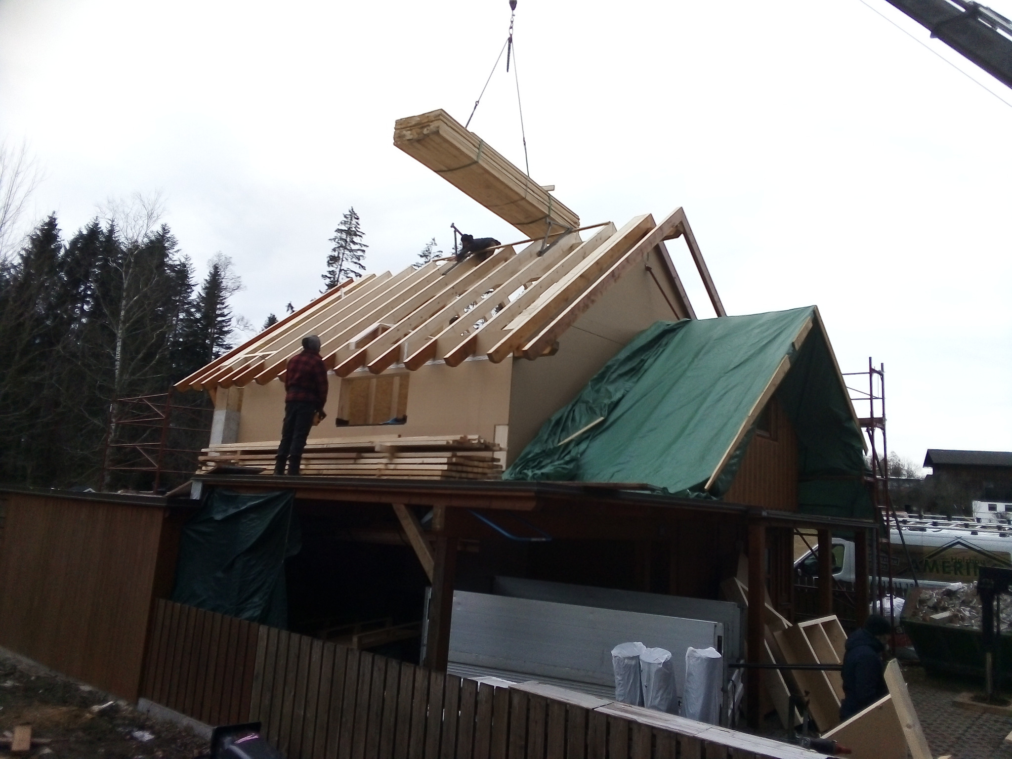 Montaż więźby dachowej: belka podnoszona dźwigiem na szkielet dachu, na tle lasu i pochmurnego nieba.