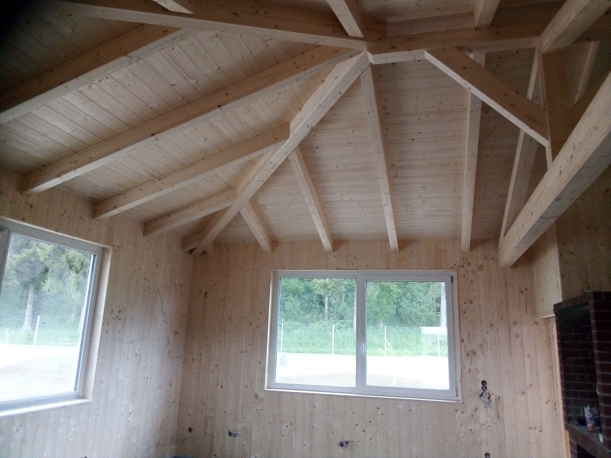 Wnętrze domu z drewnianą więźbą dachową i boazerią, widoczne okna z widokiem na zieleń.