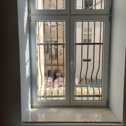 Adrian Filipczyk - Białe, dwuskrzydłowe okno z kratami w Świebodzicach, widok na podwórko z materiałami budowlanymi i rowerem, marmurowy parapet, fragment grzejnika pod oknem.