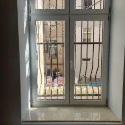 Białe, dwuskrzydłowe okno z kratami w Świebodzicach, widok na podwórko z materiałami budowlanymi i rowerem, marmurowy parapet, fragment grzejnika pod oknem.