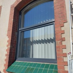 Adrian Filipczyk - Łukowe okno z szarą ramą w murze z czerwonej cegły, z zielonym ceramicznym parapetem, widoczne z boku drabiny opierającej się o ścianę.