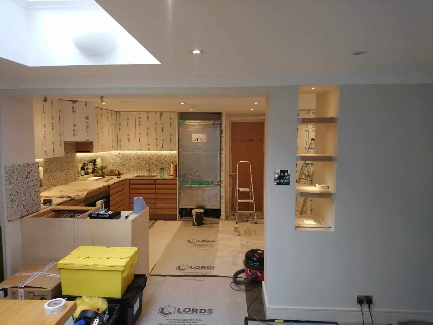 Wnętrze kuchni w trakcie montażu: widoczne zabezpieczone folią meble, przygotowane do instalacji szafki wiszące z podświetleniem, niezabudowana lodówka, kartony i narzędzia na podłodze, oraz wnęka...