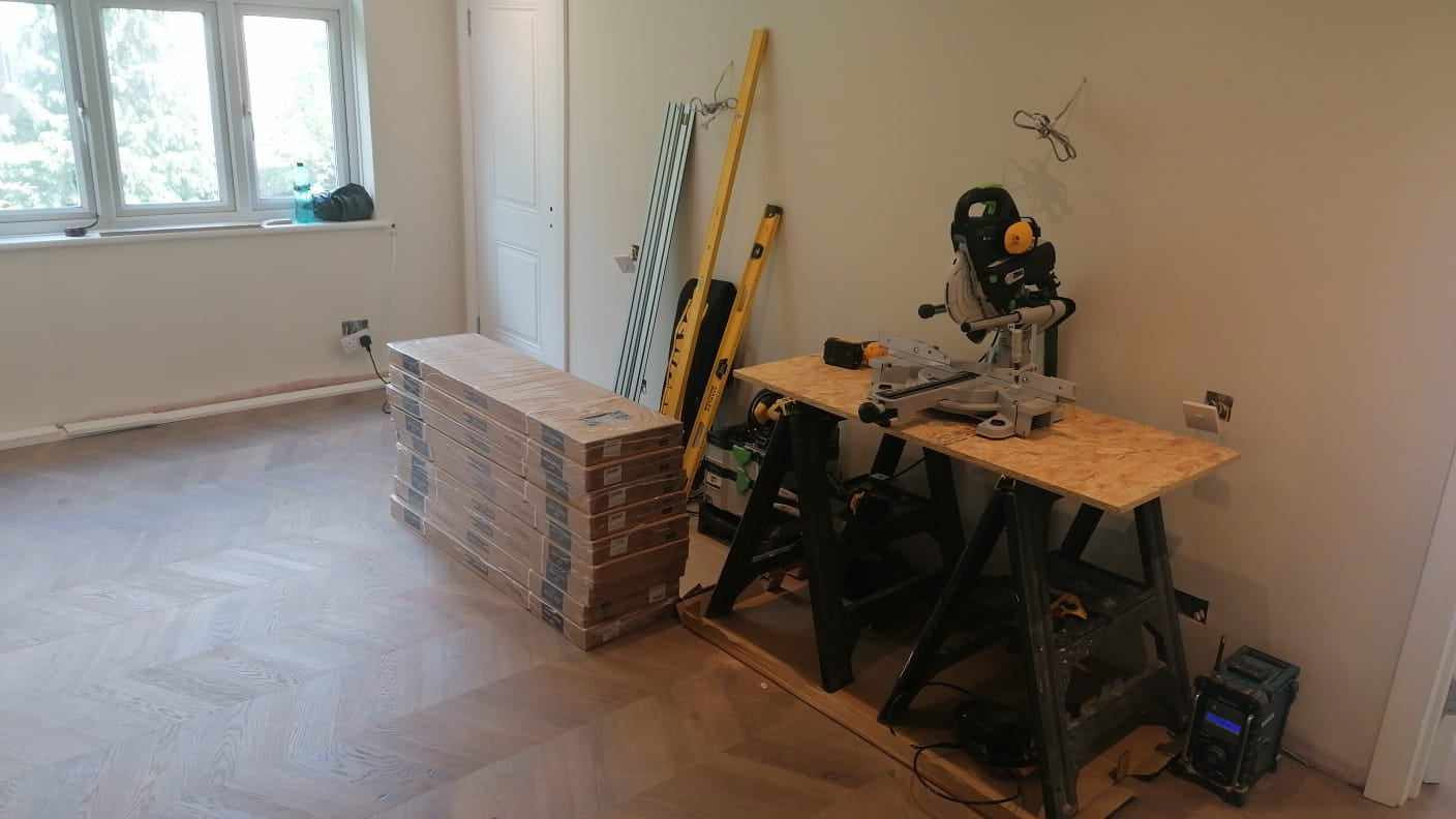 Ułożona podłoga w jodełkę francuską, paczki paneli czekają na montaż, widoczna ukośnica na stole warsztatowym i poziomice oparta o ścianę.