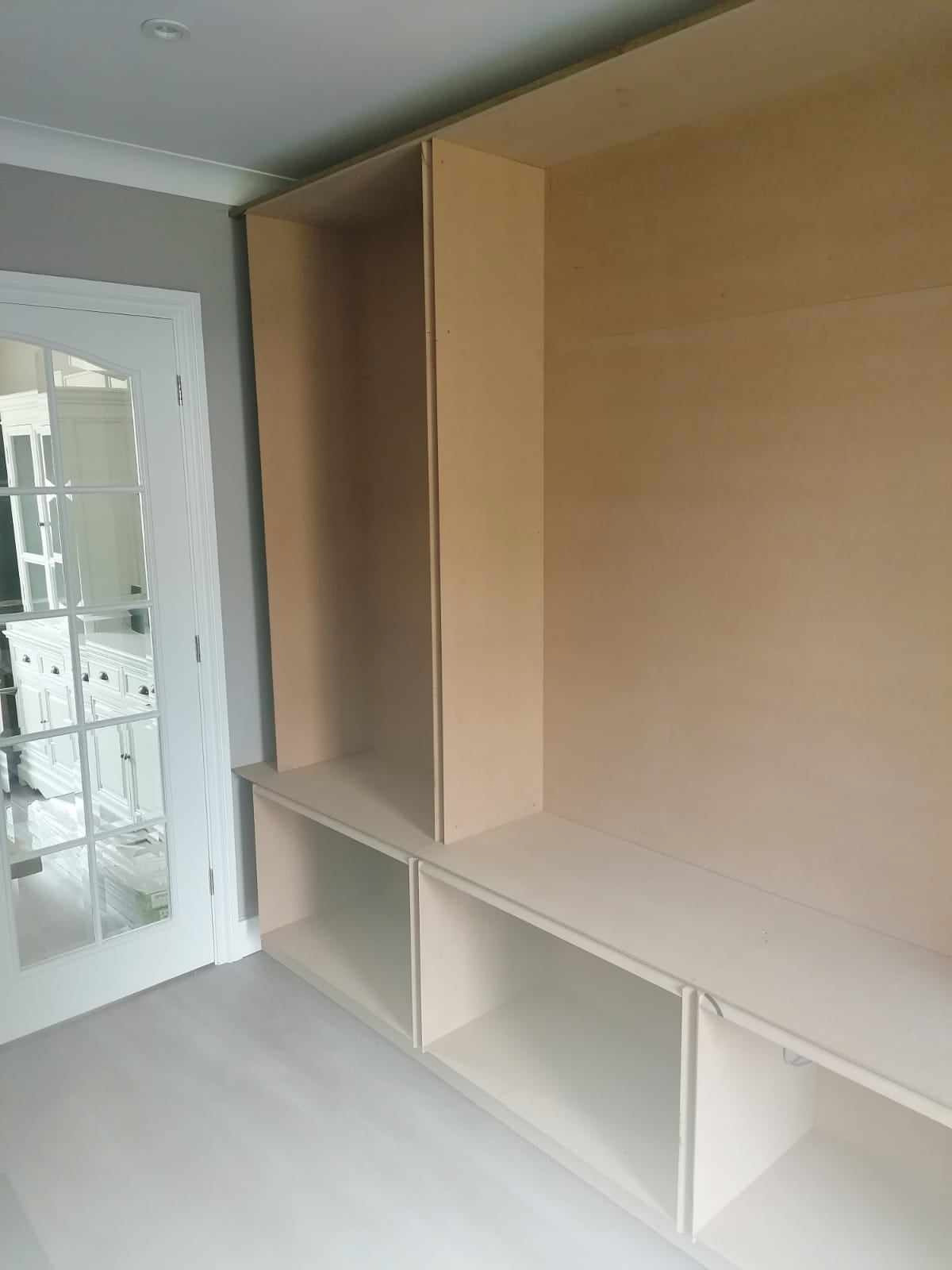 Wnętrze w trakcie tworzenia zabudowy meblowej na wymiar z surowej płyty MDF i pomalowanych na biało półek, widok na przeszklone drzwi do innego pomieszczenia.