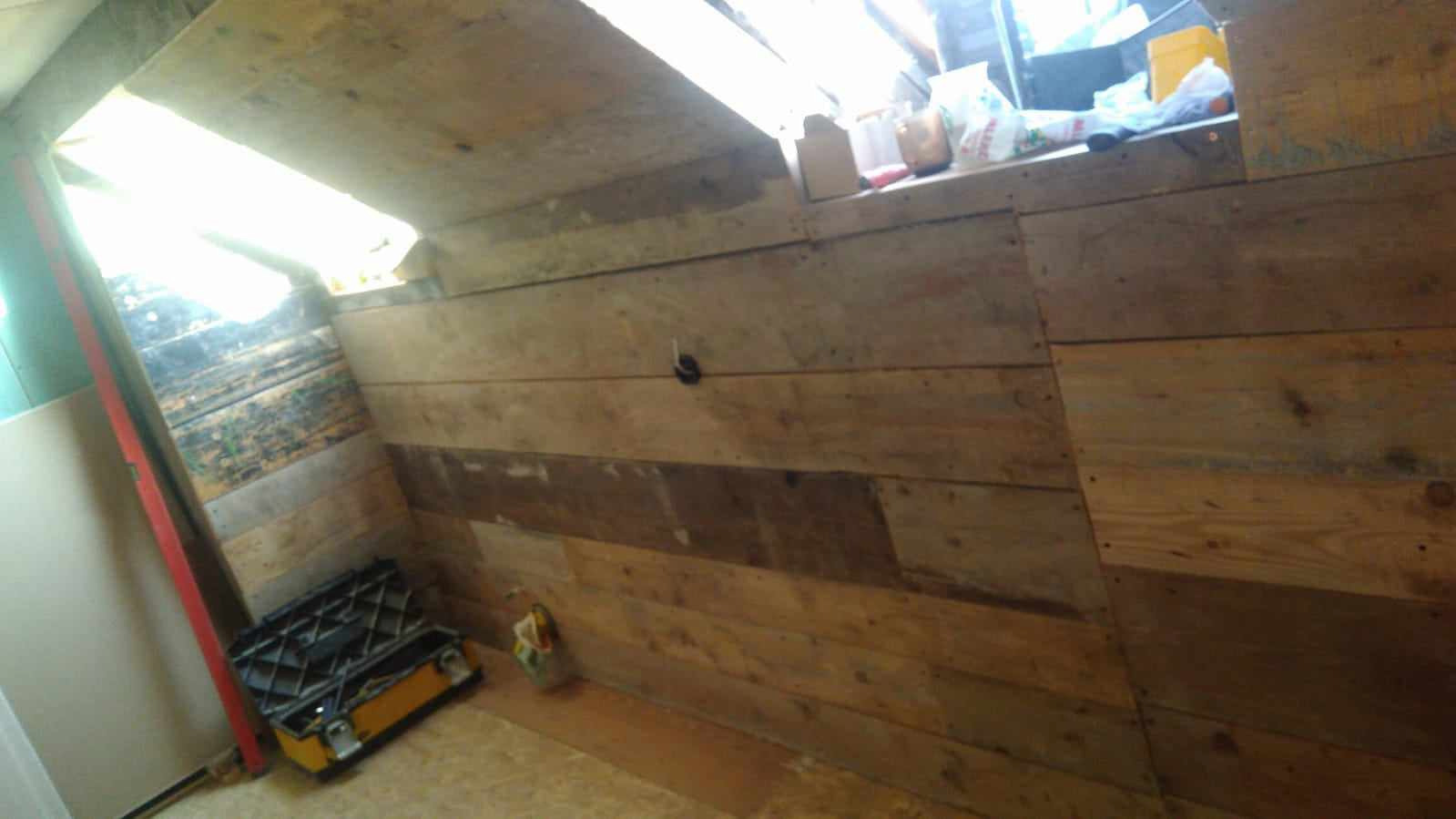 Poddasze w trakcie wykańczania drewnem, widoczna żółta skrzynka narzędziowa, poziomica oparta o ścianę i resztki materiałów budowlanych.