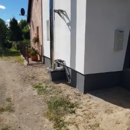 Fragment elewacji domu z zamontowanym, błyszczącym kominem stalowym z widocznymi elementami mocującymi, na tle białej ściany i piaszczystego podłoża.