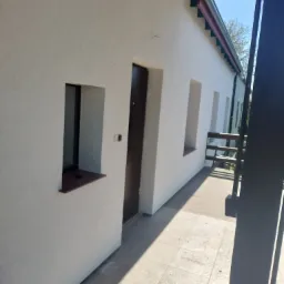 Elewacja budynku po ociepleniu i pomalowaniu na biało, widoczne okna z brązowymi parapetami i brązowe drzwi wejściowe, fragment balkonu z drewnianą podłogą.