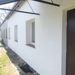 Boczny widok budynku z otynkowaną, białą elewacją, widoczne okna z brązowymi parapetami, ciemne drzwi wejściowe i fragment metalowej konstrukcji zadaszenia.