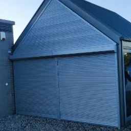 Nowoczesny budynek gospodarczy z szarymi, metalowymi drzwiami rolowanymi, otoczony żwirem.