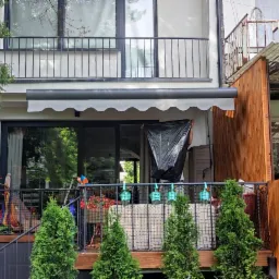 Balkon z szarą markizą z falbaną, widok z dołu na dwupiętrowy budynek, osoba w tle z czarnym workiem na balkonie.