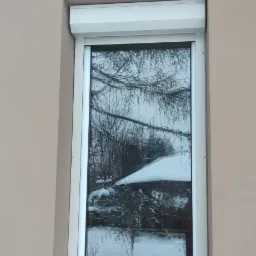 Zamknięta biała roleta zewnętrzna na oknie z widocznym odbiciem zimowego krajobrazu z zasypanym śniegiem dachem i drzewami.