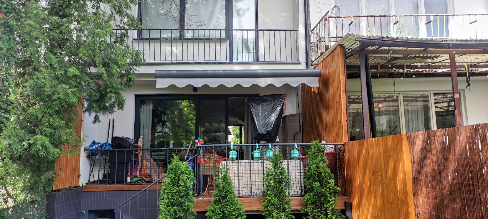 Balkon z szarą markizą z falbaną, widok z dołu na dwupiętrowy budynek, osoba w tle z czarnym workiem na balkonie.