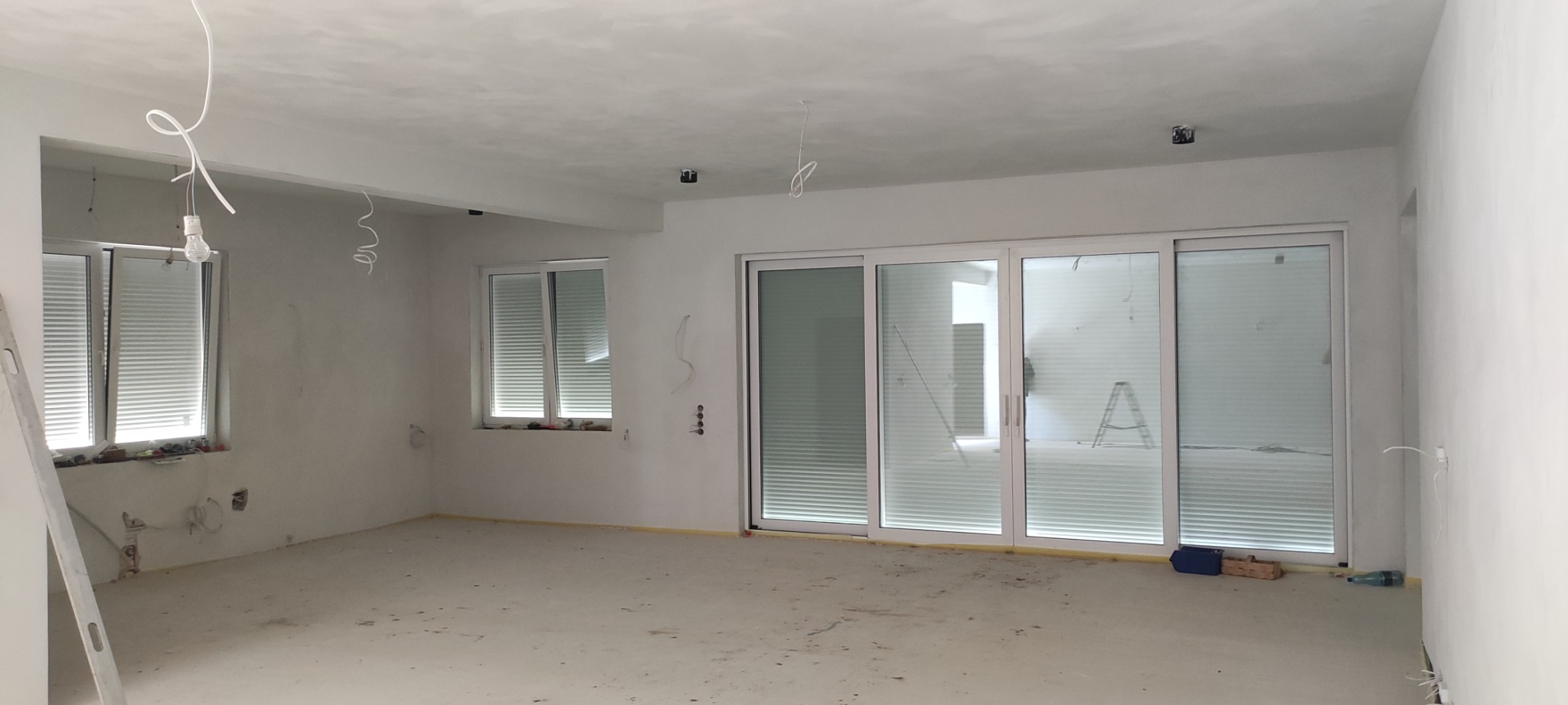 Surowe wnętrze w trakcie wykańczania z widocznymi oknami z wewnętrznymi żaluzjami, przesuwne okno tarasowe, wystające kable i drabina w tle.