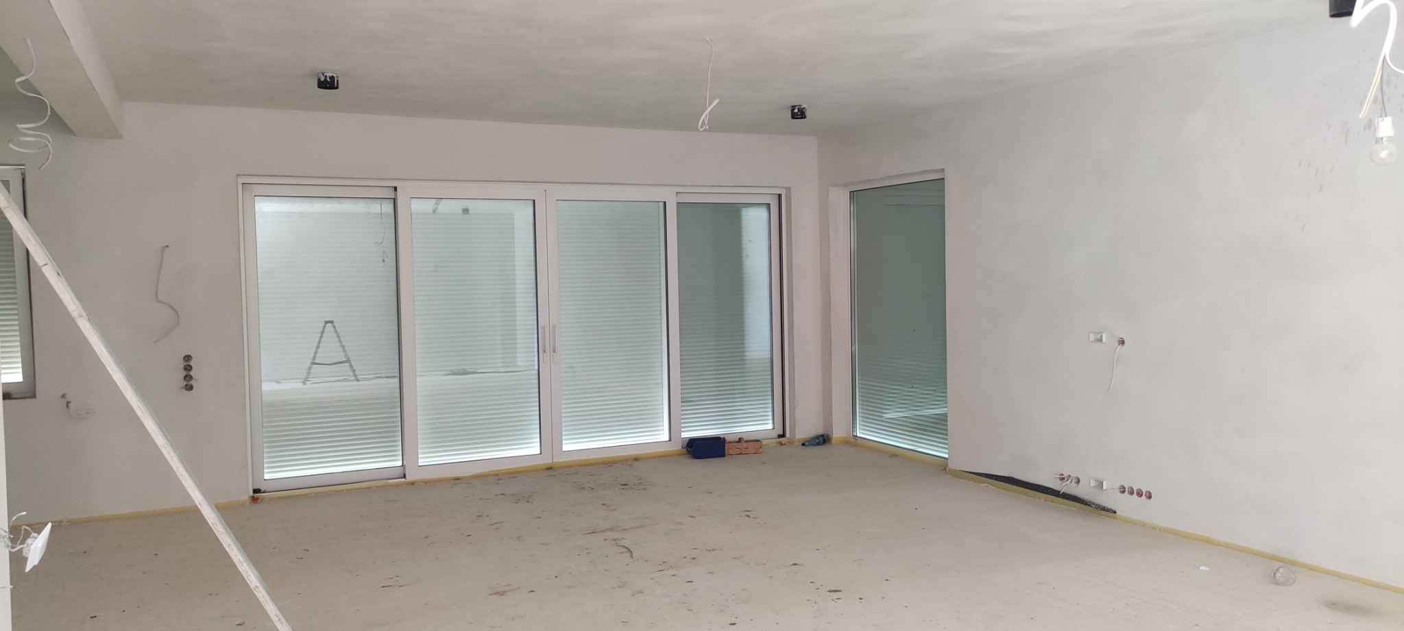 Wnętrze pokoju w trakcie remontu z widocznymi oknami z zamontowanymi żaluzjami wewnętrznymi, drabina oparta o ścianę, widoczne puszki elektryczne bez osprzętu.