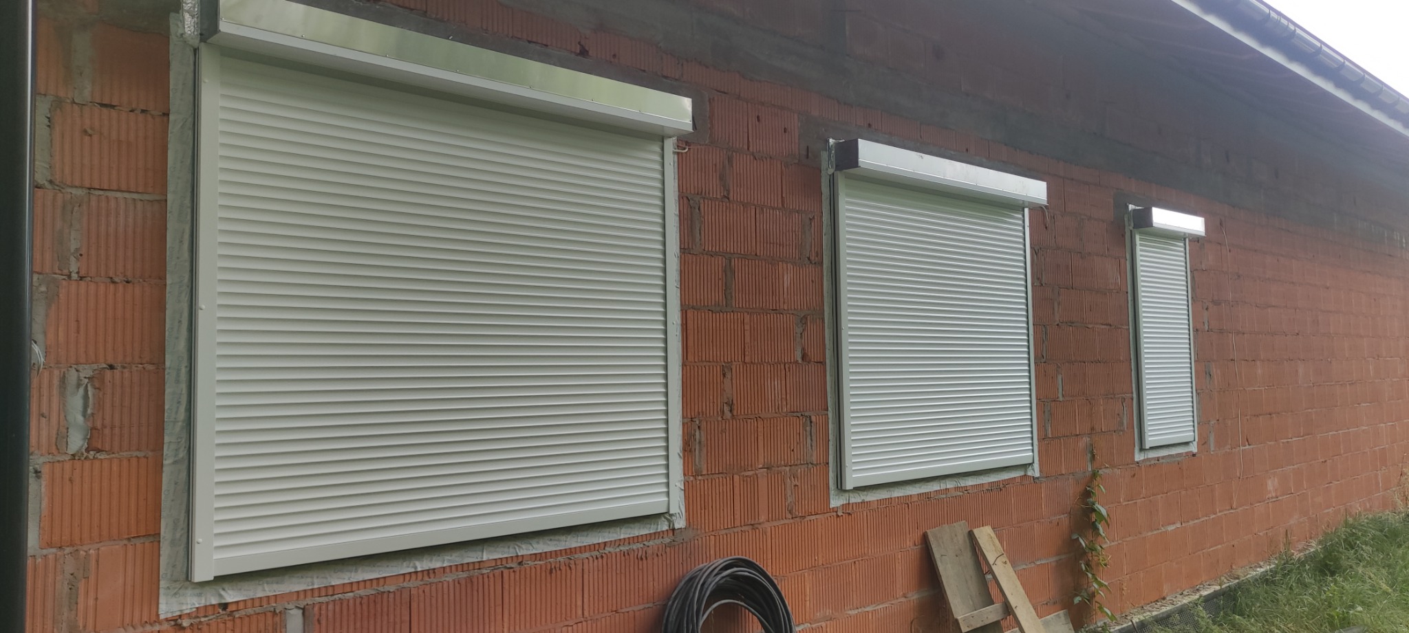 Trzy okna z zamontowanymi, opuszczonymi, jasnymi osłonami zewnętrznymi na surowej, ceglanej ścianie budynku w trakcie budowy.