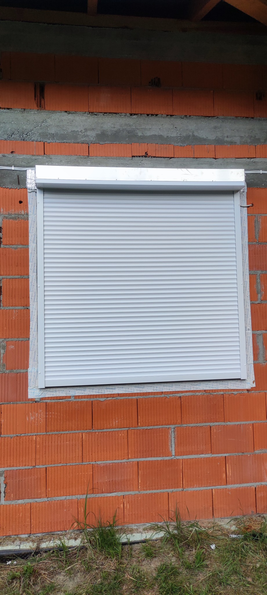 Zamknięta szara osłona okienna zamontowana w surowej, ceglanej ścianie budynku w trakcie budowy.