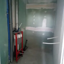 Wnętrze łazienki w trakcie prac, widoczne rury, stelaż podtynkowy WC na tle surowych ścian z płytami gipsowo-kartonowymi, z widocznymi łączeniami i śladami szpachlowania.