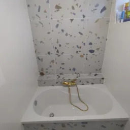 Biała wanna obudowana płytkami z wzorem lastryko, złota bateria natryskowa, ściany wyłożone płytkami o podobnym wzorze, wnęka z kosmetykami.