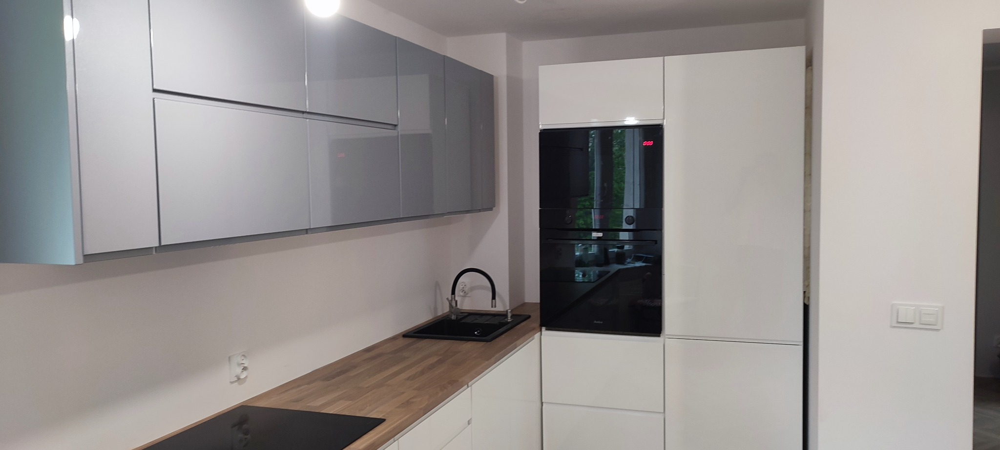 Nowoczesna kuchnia z szarymi szafkami wiszącymi, drewnianym blatem, czarnym zlewem i piekarnikiem w zabudowie.