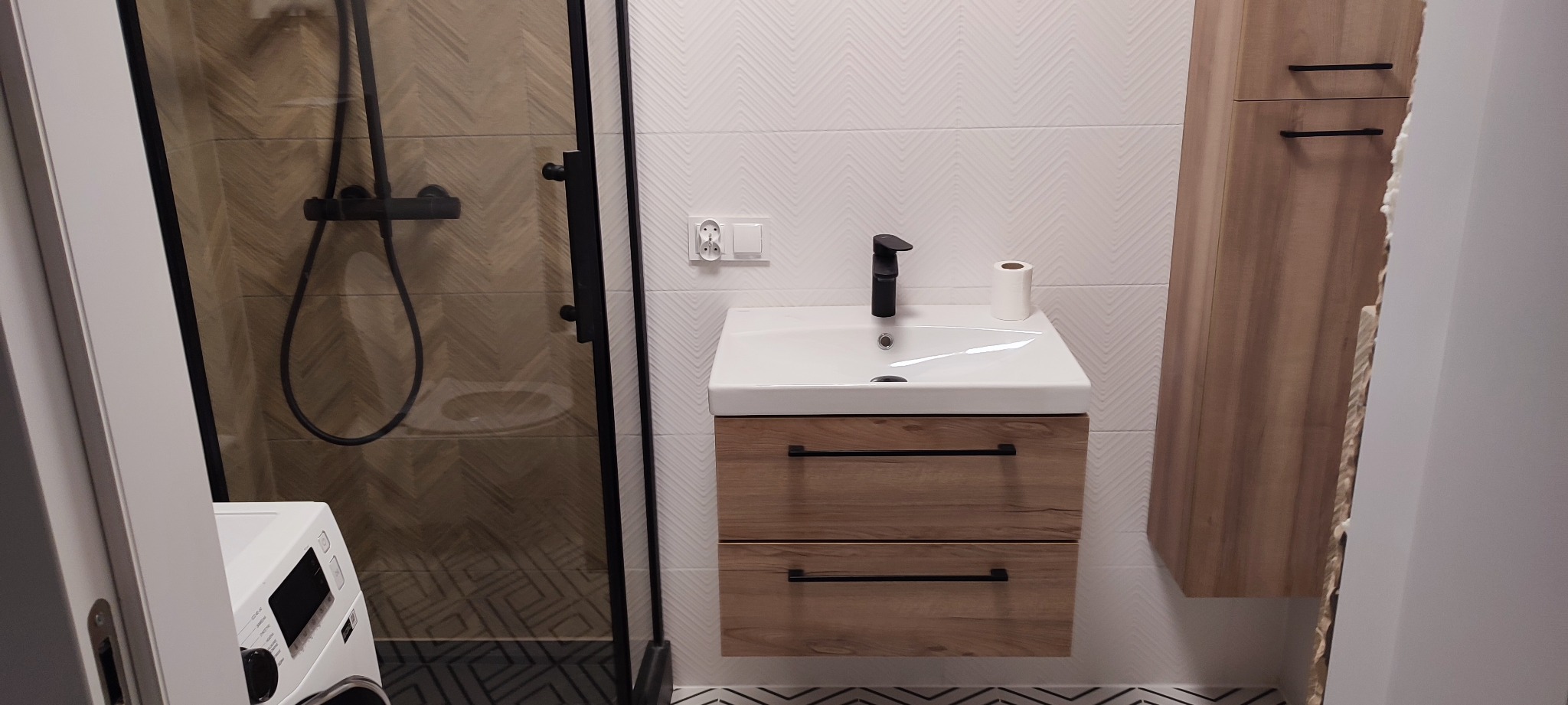 Nowoczesna łazienka z kabiną prysznicową z czarnymi elementami, umywalką z szafką w kolorze drewna i wiszącą szafką, wykończona białymi płytkami w jodełkę.