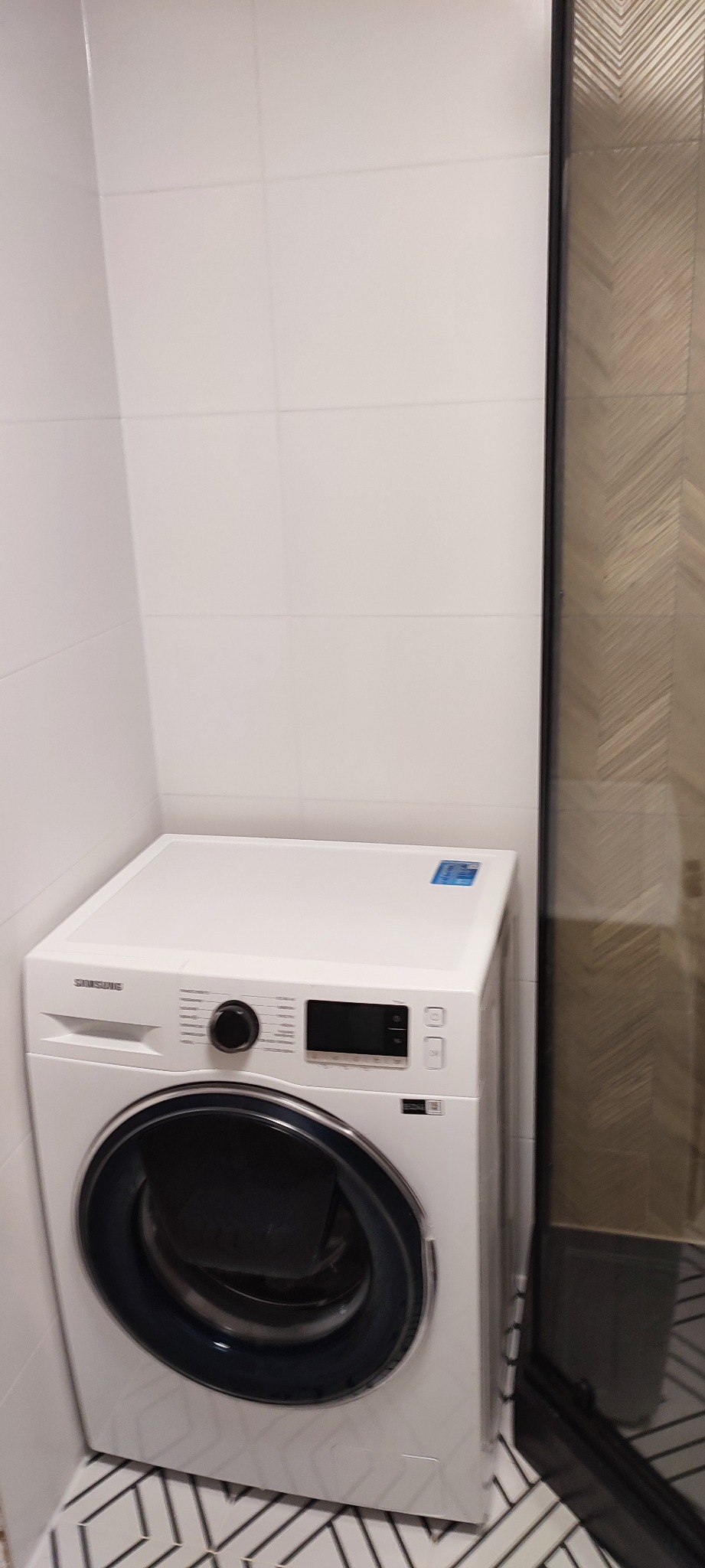 Nowoczesna pralka Samsung w białej łazience z geometryczną posadzką i częściowo widoczną kabiną prysznicową z wzorzystą ścianą.