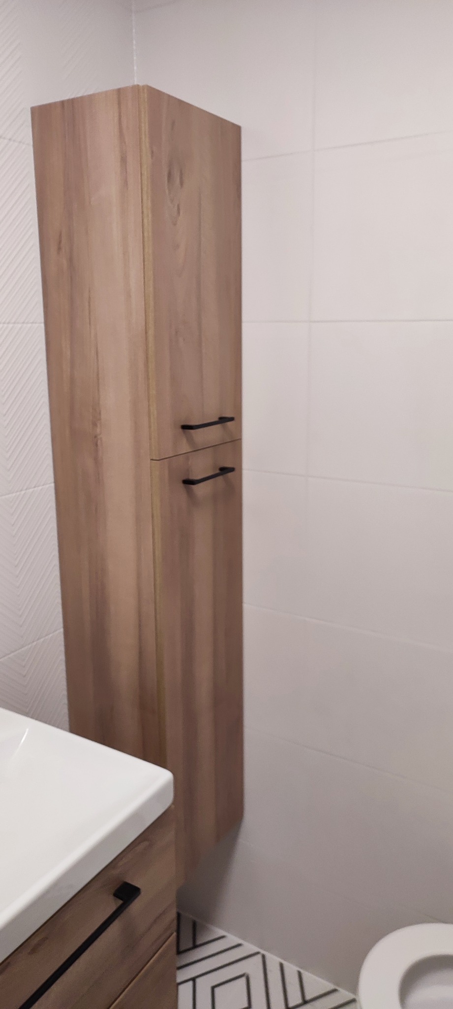 Wysoka, drewnopodobna szafka łazienkowa z dwoma szufladami i czarnymi uchwytami, zamontowana na tle białych płytek w łazience, widoczny fragment umywalki i toalety.
