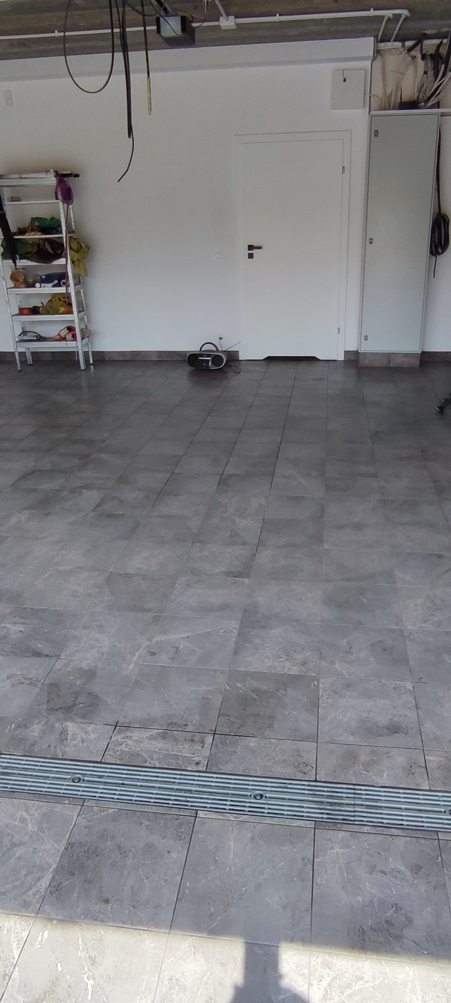 Szare płytki podłogowe imitujące marmur, z metalowym odpływem liniowym w garażu lub pomieszczeniu gospodarczym. W tle białe ściany, drzwi i regał.