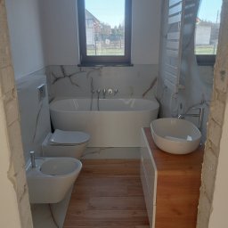 WZ Dekor - Nowoczesna łazienka po remoncie z wanną, bidetem, umywalką nablatową na drewnianej szafce i grzejnikiem drabinkowym, wykończona płytkami imitującymi marmur i drewno, widok przez wejście w trakcie...
