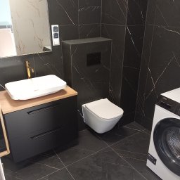 WZ Dekor - Nowoczesna łazienka z czarnymi płytkami imitującymi marmur, białą umywalką nablatową z złotą armaturą, podwieszaną toaletą i pralką.