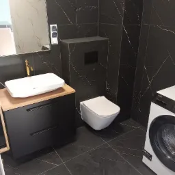 Nowoczesna łazienka z czarnymi płytkami imitującymi marmur, białą umywalką nablatową z złotą armaturą, podwieszaną toaletą i pralką.