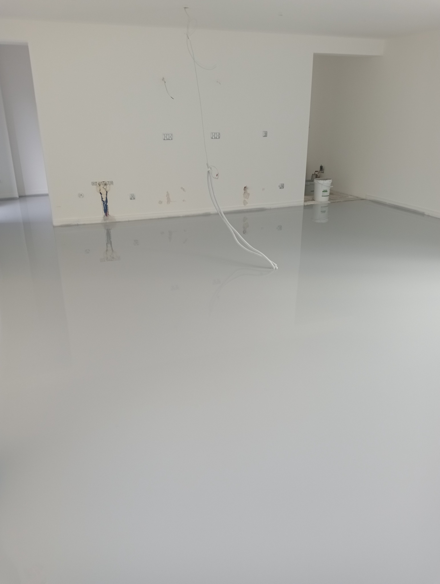 Jasnoszara, błyszcząca posadzka w nowym, pustym pomieszczeniu z białymi ścianami, widoczne puszki elektryczne i wystające kable, w tle wiadro z farbą.