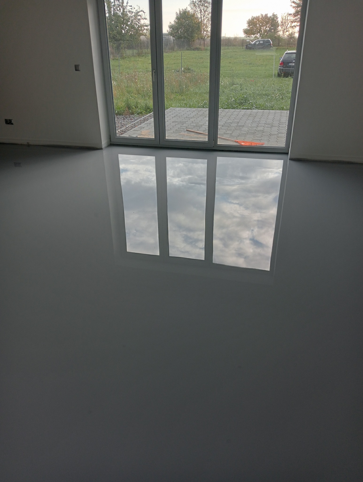 Błyszcząca, szara podłoga w nowym wnętrzu odbija okno tarasowe z widokiem na ogród z zaparkowanymi samochodami.