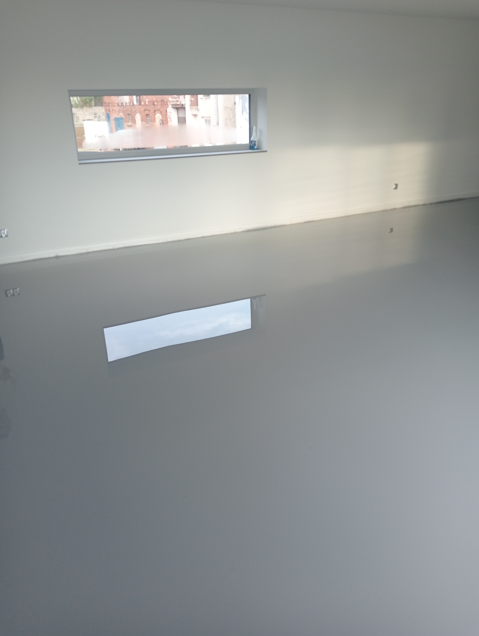 Szara, gładka posadzka w pustym pomieszczeniu z oknem, w którym odbija się budynek z cegły.