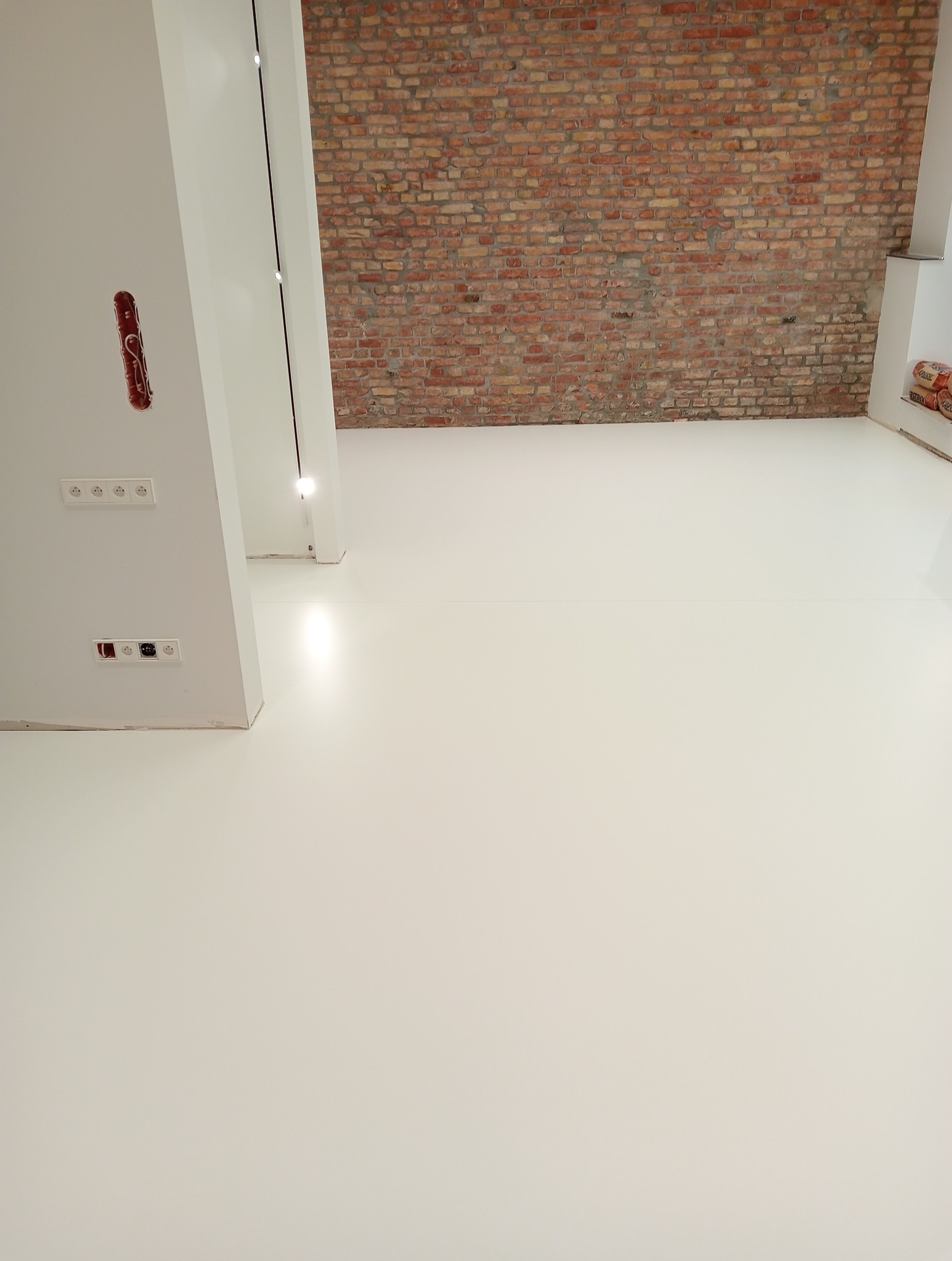 Biała, jednolita podłoga w jasnym pomieszczeniu z odsłoniętą ceglaną ścianą i widocznymi gniazdkami elektrycznymi w ścianie.