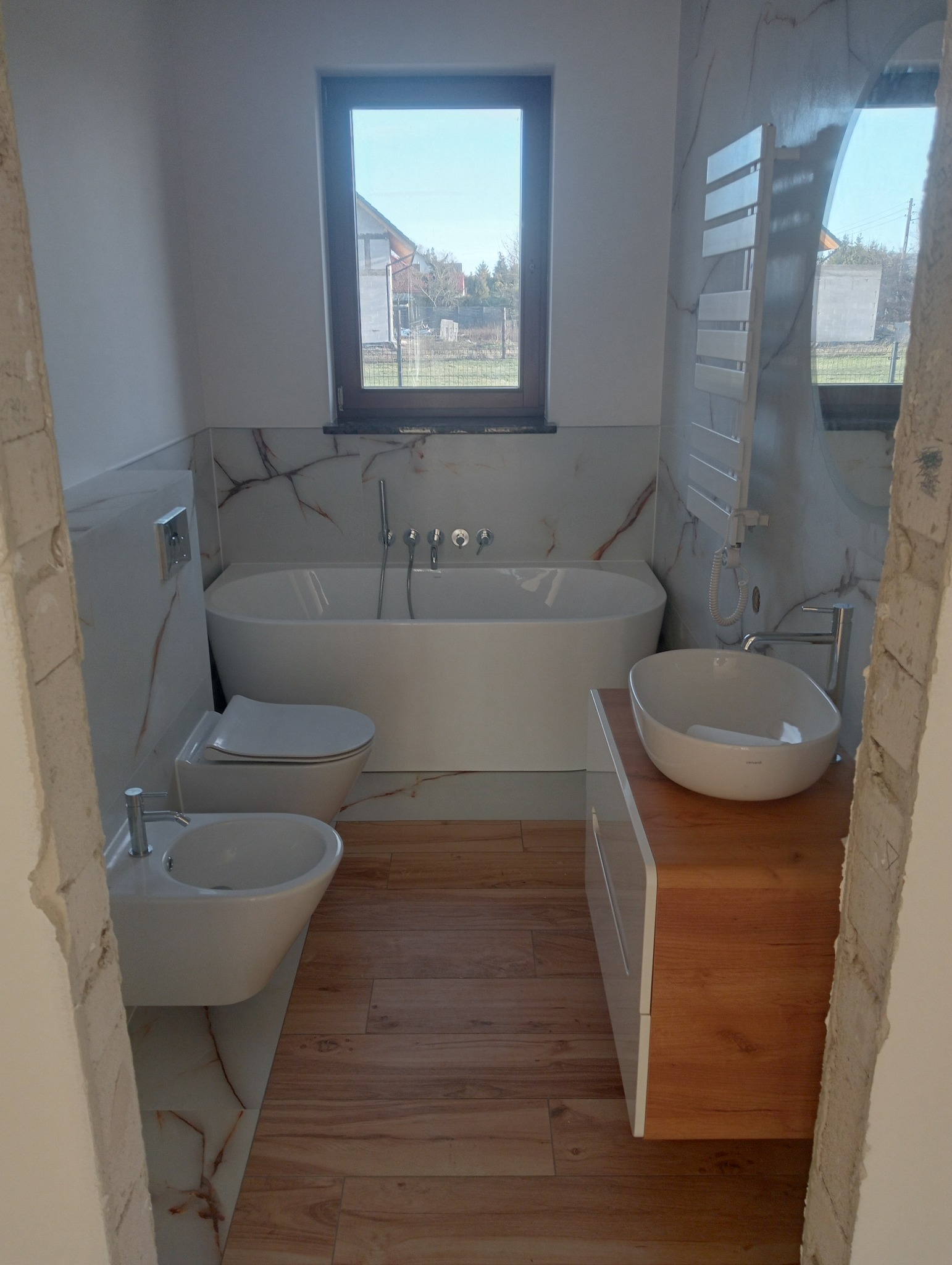 Nowoczesna łazienka po remoncie z wanną, bidetem, umywalką nablatową na drewnianej szafce i grzejnikiem drabinkowym, wykończona płytkami imitującymi marmur i drewno, widok przez wejście w trakcie...