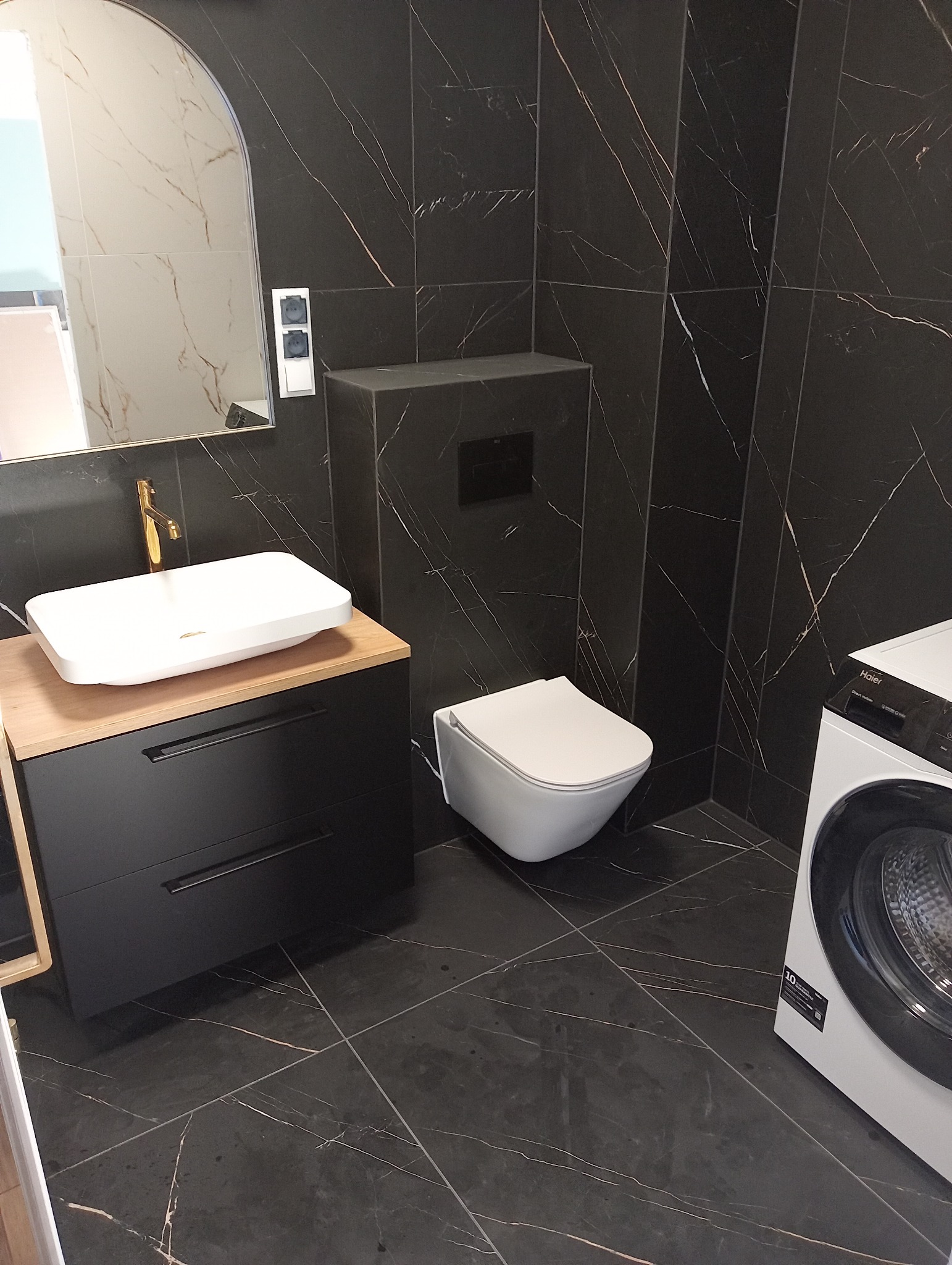 Nowoczesna łazienka z czarnymi płytkami imitującymi marmur, białą umywalką nablatową z złotą armaturą, podwieszaną toaletą i pralką.