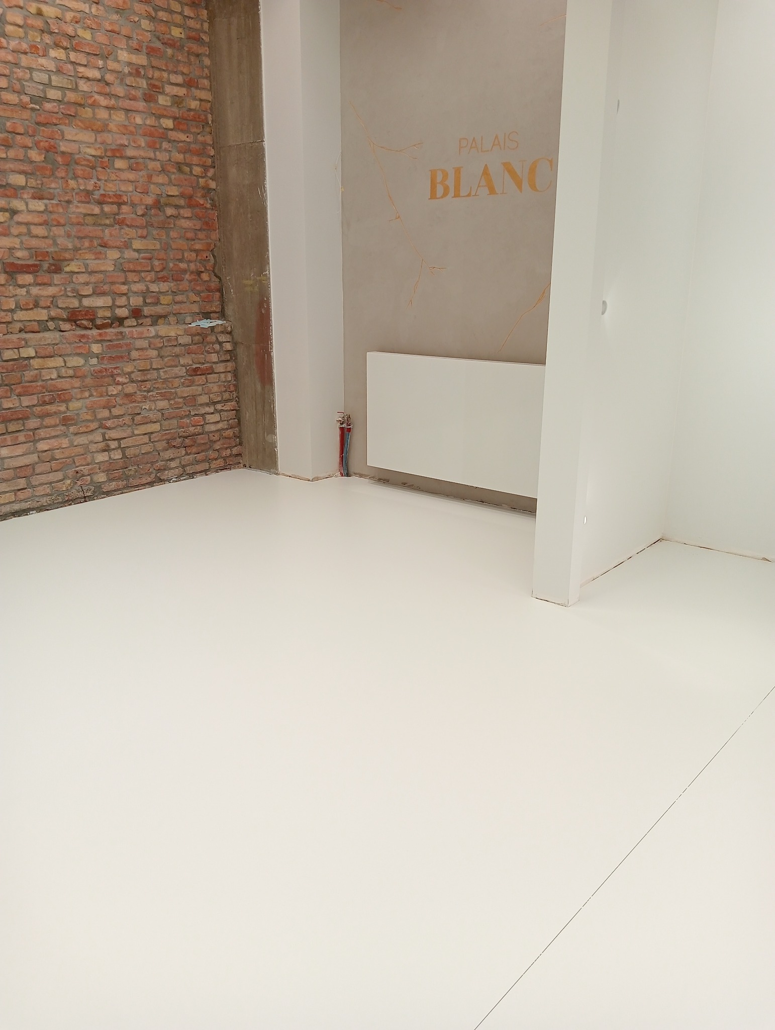 Biała posadzka żywiczna w surowym wnętrzu z odsłoniętą cegłą i fragmentem ściany z napisem 'Palais Blanc'.