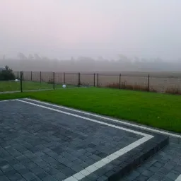Ułożona kostka brukowa w odcieniach szarości z białymi akcentami obramowania, przylegająca do zadbanego trawnika z czarnym ogrodzeniem w tle, spowitego poranną mgłą.