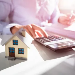Model domu obok kalkulatora i dokumentów, osoba w tle pisze długopisem, jasne oświetlenie.