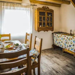 Jasna kuchnia w stylu rustykalnym z drewnianym stołem nakrytym do posiłku, drewnianymi krzesłami z poduszkami w kwiaty,  wiszącą szafką i zlewem z blatem zakrytym kwiecistą tkaniną.