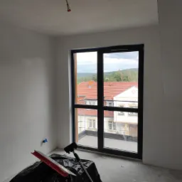 Pokój w trakcie wykańczania: ściany po nałożeniu gładzi, widoczne narzędzia do gładziowania zabezpieczone folią, okno z widokiem na budynki z czerwonymi dachami.