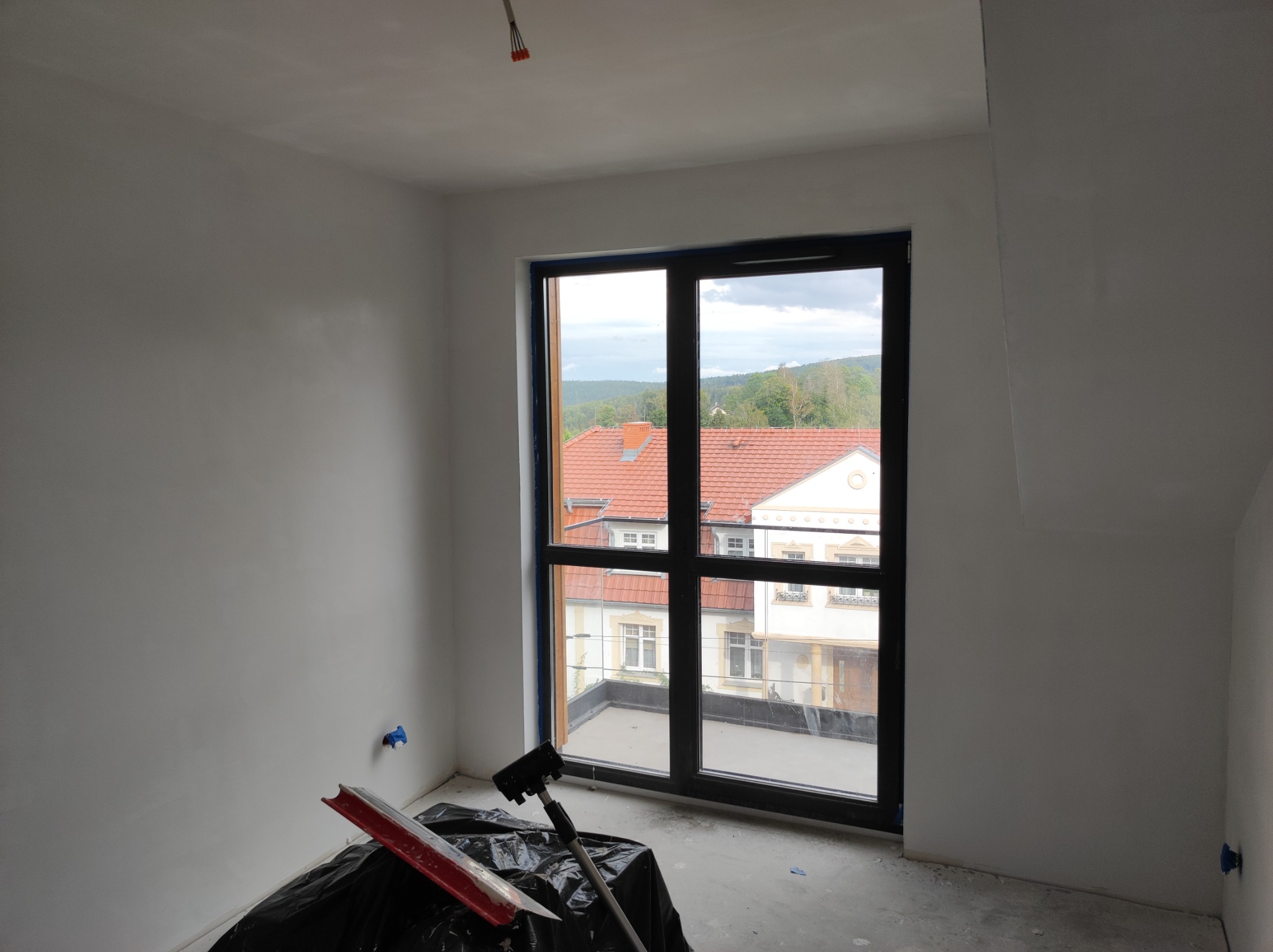 Pokój w trakcie wykańczania: ściany po nałożeniu gładzi, widoczne narzędzia do gładziowania zabezpieczone folią, okno z widokiem na budynki z czerwonymi dachami.