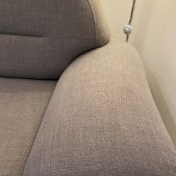 WrapStudio - Mateusz Kędzior - Fragment szarej, tapicerowanej kanapy z widocznym podłokietnikiem i oparciem, oświetlony lampą stojącą w tle.