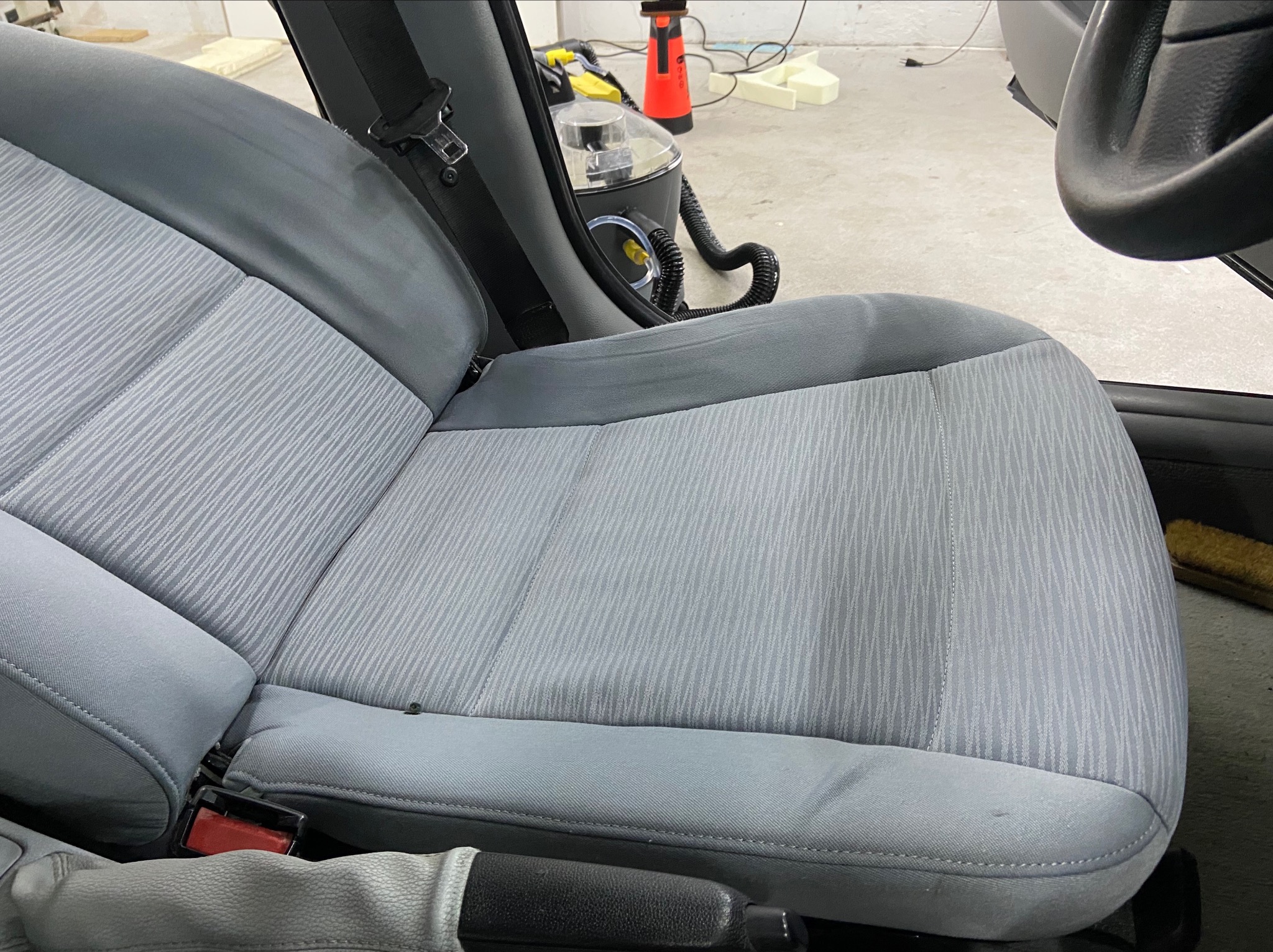 Szare, materiałowe siedzenie samochodowe z widocznym pasem bezpieczeństwa i fragmentem kierownicy, obok stoi odkurzacz piorący z wężem.
